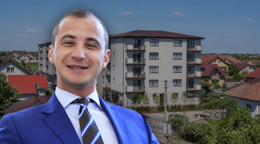 Smiling man in a blue suit and striped tie with a residential building behind him.