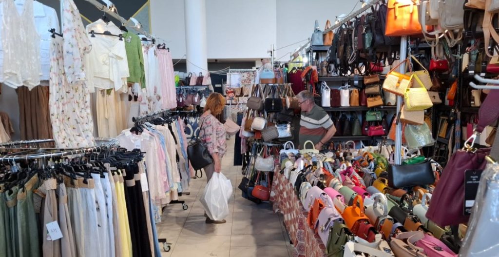 siu 4 - Sursa de Vest Two shoppers browse a crowded handbag stall with shelves full of purses on both sides in a market setting.