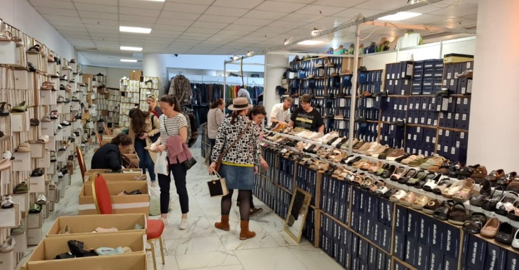 siu 6 - Sursa de Vest Group of shoppers in a shoe store browsing rows of shoes and boxes along shelves.