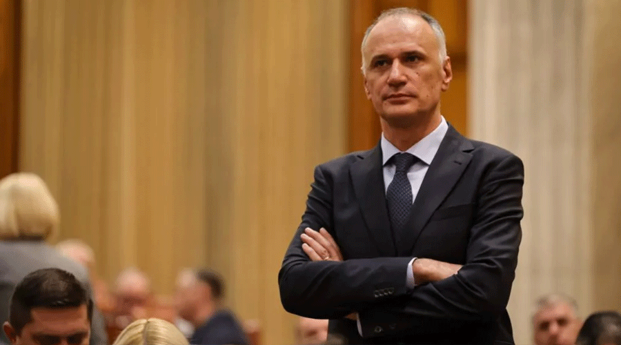 Businessman in a dark suit stands with arms crossed at a formal event, blurred attendees in the background.