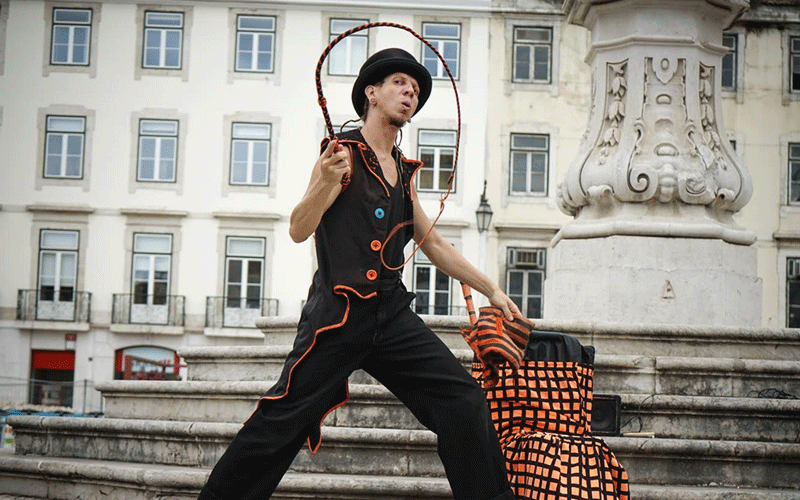 Street performer in a black outfit with orange trim spinning a hoop in front of a historic square and an ornate stone pedestal.