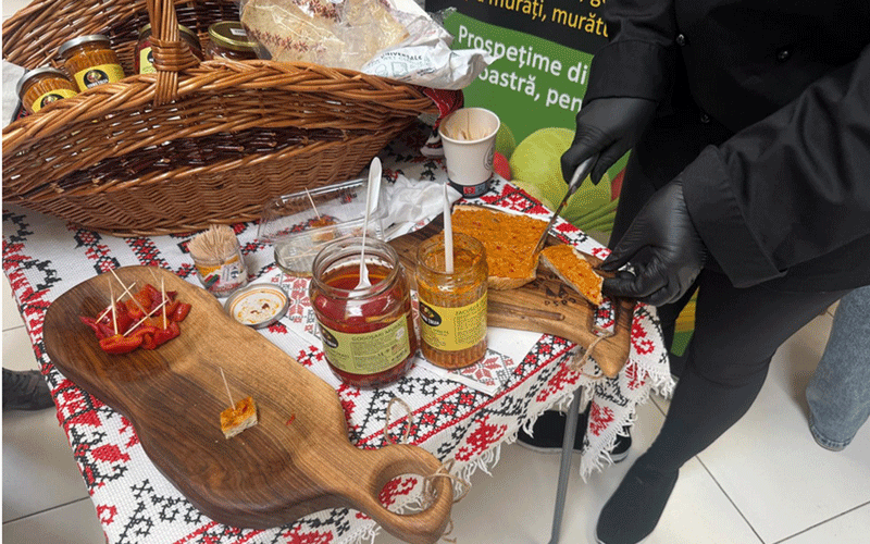 Chef in black gloves slicing bread at a tasting table with jars of jam and peppers on a patterned cloth