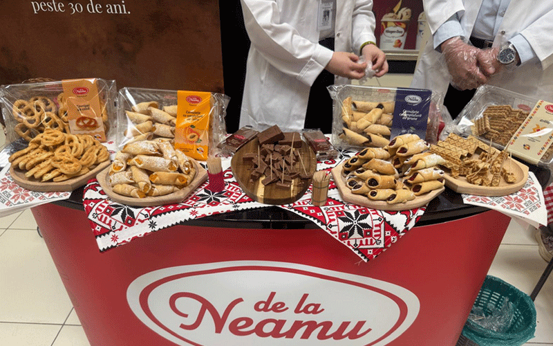 Assorted cookies and pastries on plates arranged on a red De la Neamu display table with two people in white coats.