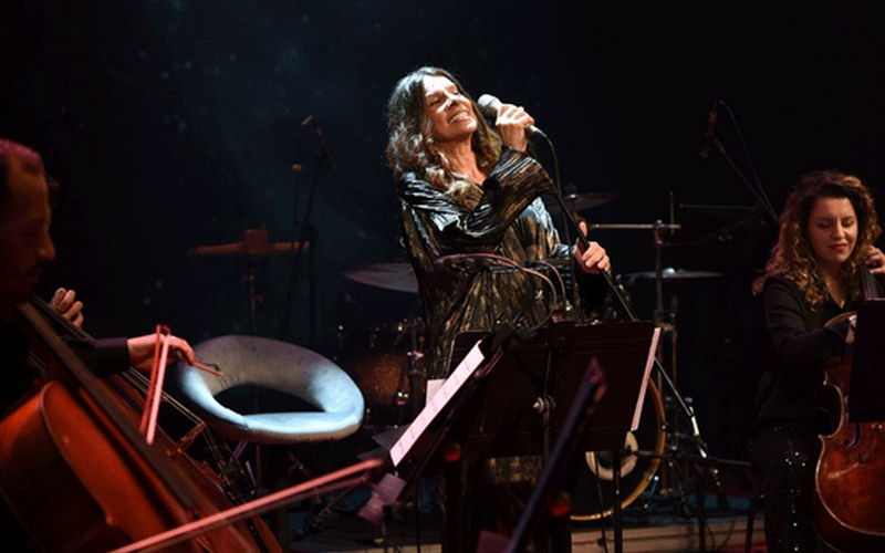Female vocalist singing into a microphone on stage while a cellist and another string player accompany in the background on a dimly lit concert set.