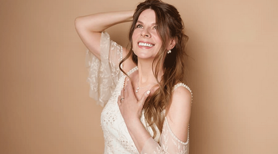 Bride in a white lace wedding dress, smiling with one hand on her chest against a beige backdrop.