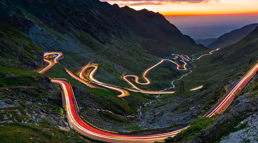 Long-exposure shot of a winding mountain road at dusk, with red and white light trails from passing cars in a valley.