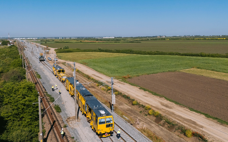 Maintenance train on a rural railway line with workers nearby and farmland on both sides under a clear blue sky