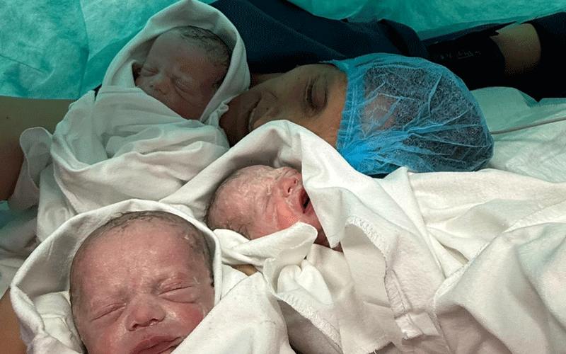 Mother resting in a hospital bed with two newborn twins swaddled in white blankets beside her.