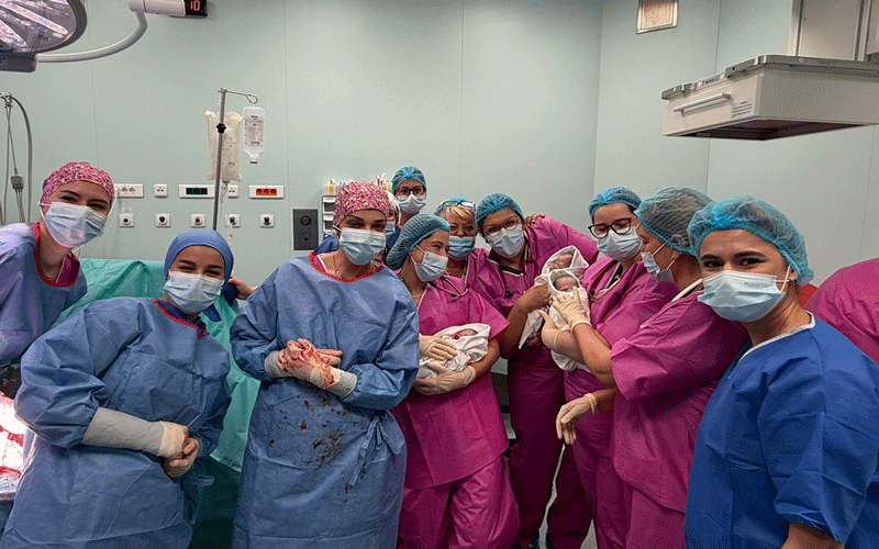 Surgical team in blue and pink gowns, masks, and caps gathers around a newborn baby in an operating room.