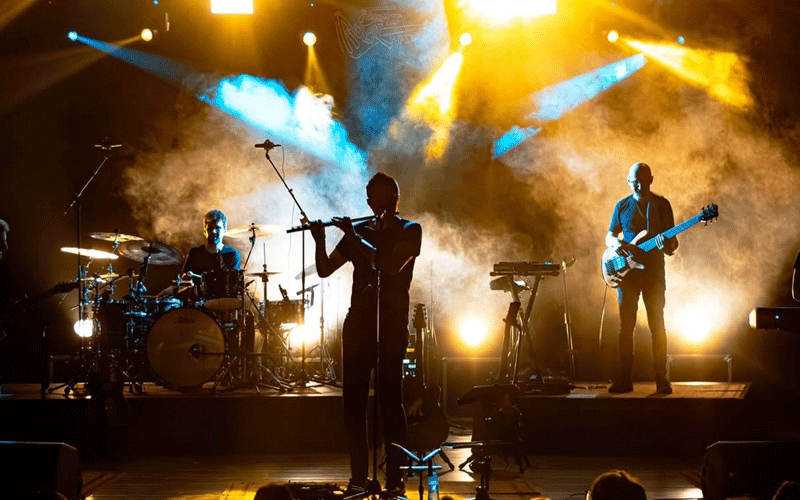 A three-person band performs on a smoky stage: drummer left, flute player center, bassist right, under blue and yellow stage lights.
