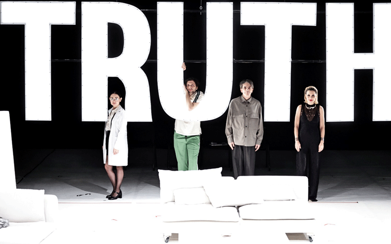Four performers stand on a stage behind oversized white letters spelling ‘TRUTH’, with a white sofa and pillows in the foreground.