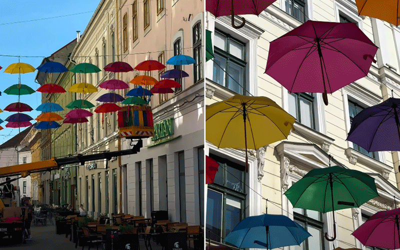 Colorful umbrellas suspended above a sunny street with outdoor café seating and pastel buildings on either side.