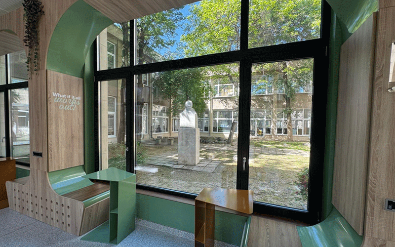 Indoor seating nook with wooden and green furniture beside a large glass wall; outside is a courtyard with trees and a statue.