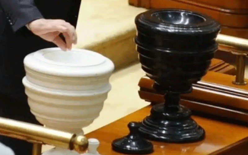 White communion chalice and a black chalice on a wooden altar rail in a church setting, with a hand near the white chalice