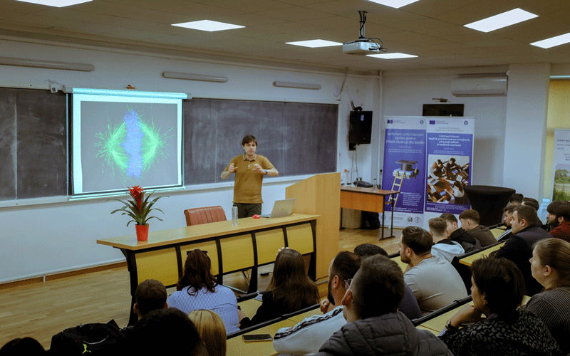 Lecturer speaks at a wooden podium in a university classroom; projection screen shows a colorful abstract image, while students listen.