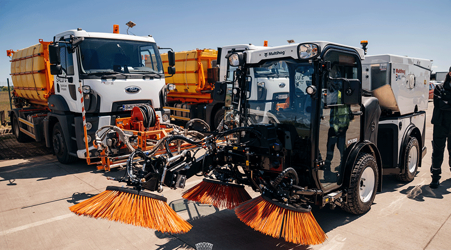 Street sweeper with large orange rotating brushes parked beside an orange refuse truck on a sunny day