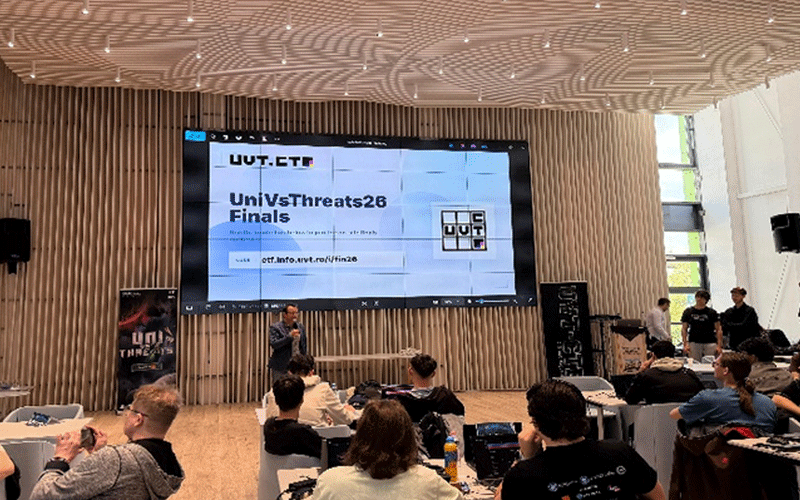Speaker on stage at UniVsThreats26 Finals with a large screen displaying the event title and logo behind him, in a modern wooden-walled auditorium.
