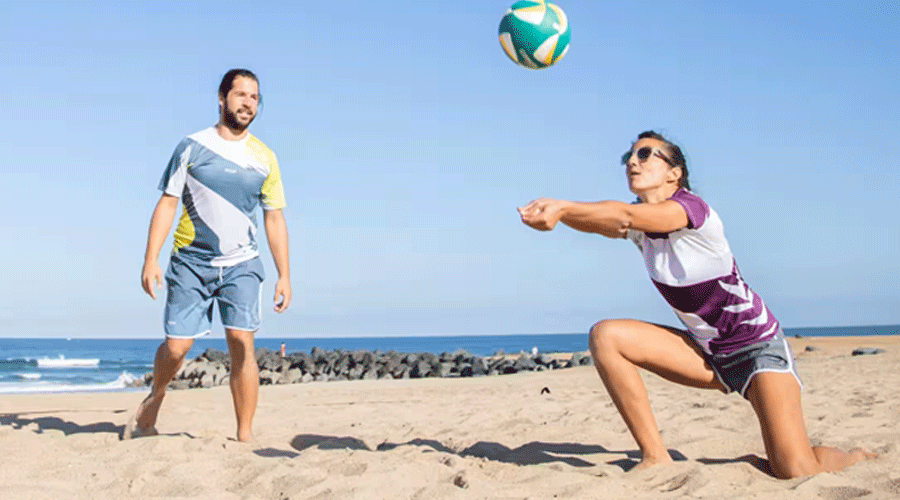 Two beach volleyball players on sandy shore; a man stands left while a woman dives to hit a ball in midair with the ocean in the background.