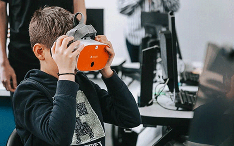 Boy wearing an orange virtual reality headset at a computer station in a classroom tech setup