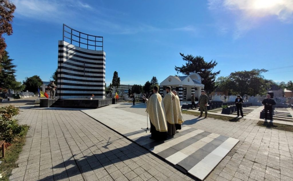 ziua eroilor 1 - Sursa de Vest Three clergy in gold robes lead a ceremony beside a tall curved black-and-white striped memorial in a plaza, with soldiers and attendees nearby on a sunny day.