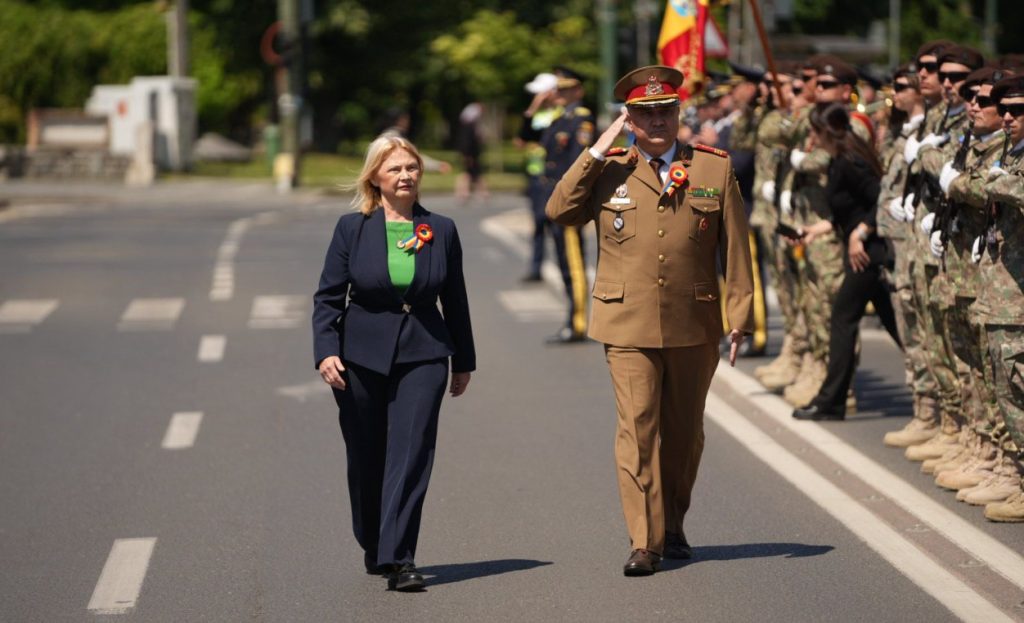 ziua eroilor 3 - Sursa de Vest A woman in a navy suit walks beside a saluting military officer on a parade street with uniformed soldiers in formation behind them.