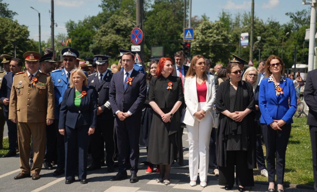 ziua eroilor 4 - Sursa de Vest Group of officials and civilians standing on a street during a ceremony; several in military uniforms and suits, all wearing orange-red rosettes.