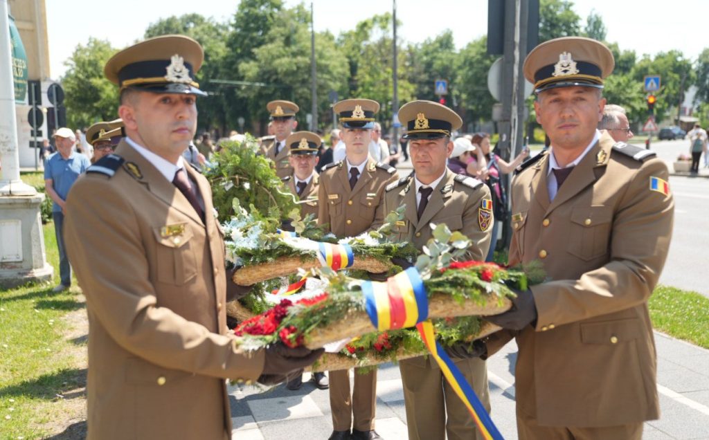 ziua eroilor 7 - Sursa de Vest Military officers in tan uniforms carry a flower wreath tied with blue, yellow, and red ribbons during a ceremony outside on a sunny day
