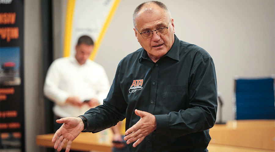 Middle-aged man in a dark shirt with AJTA logo gesturing while speaking in a workshop, with a blurred assistant in the background.