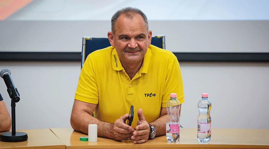 Man in a bright yellow polo speaks at a panel, seated at a wooden table with a microphone and water bottles in front of him