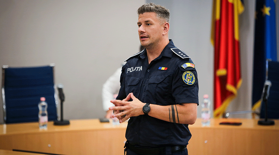 Policeman in a dark blue police uniform speaking in a conference room with flags in the background and a table in front.