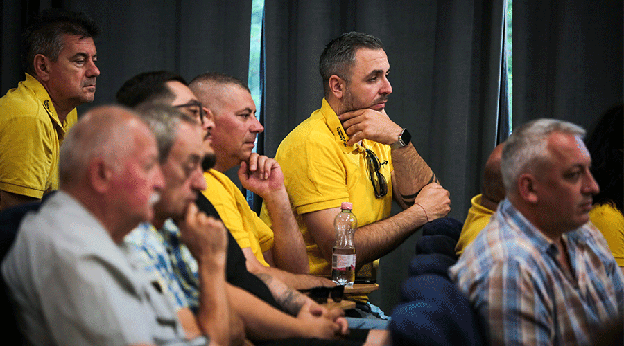 Group of men in yellow shirts seated indoors, attentively listening during a meeting conference or briefing.