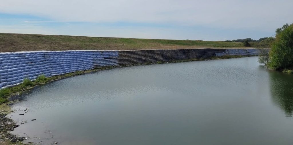 aba banat - raul timis 2 - Sursa de Vest A canal with a steep embankment reinforced by blue sandbags stacked in a protective wall along the water surface and a grassy slope beyond edge.