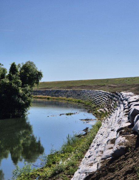 aba banat - raul timis 3 - Sursa de Vest Curved stone embankment along a calm pond, with a green hill and blue sky, and a tree reflected in the water.