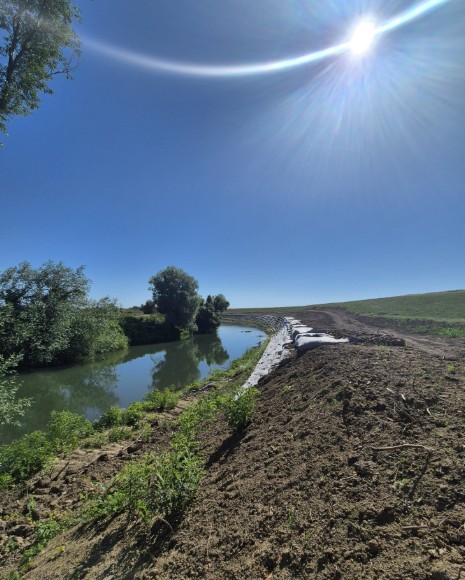 aba banat - raul timis 4 - Sursa de Vest Bright sunny day along a narrow river, with trees on the left and a dirt embankment and path on the right extending toward the horizon.