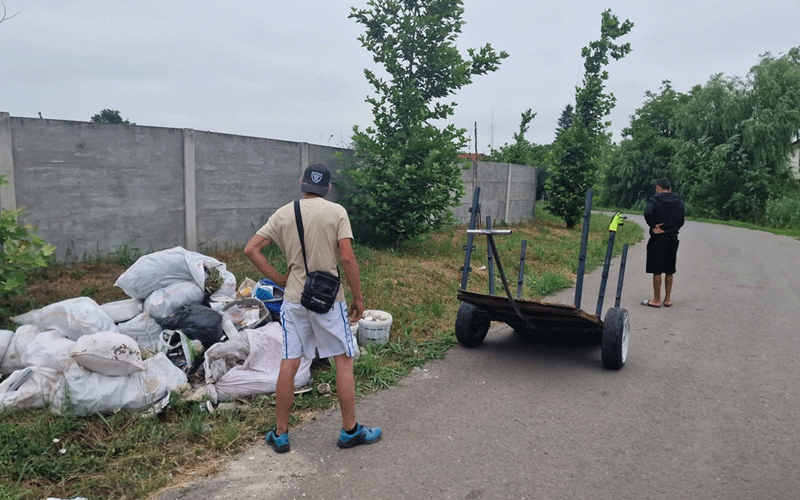 Two men conduct a roadside cleanup: one stands near a pile of trash bags beside a concrete wall, while a wheeled cart sits on the road and another man watches from a distance.