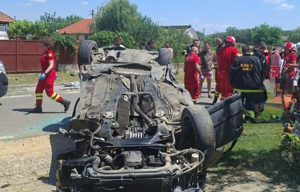 Upside-down car on a sunny street, surrounded by emergency responders and firefighters assessing the wreckage.