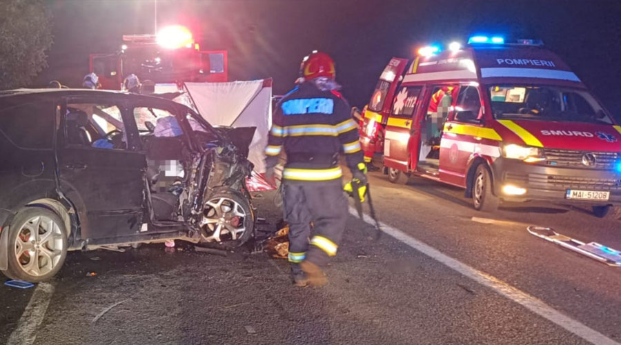 accident-bh1 - Sursa de Vest Nighttime highway crash scene with a severely damaged black car, a firefighter in gear walking away, and an ambulance with flashing lights in the background.