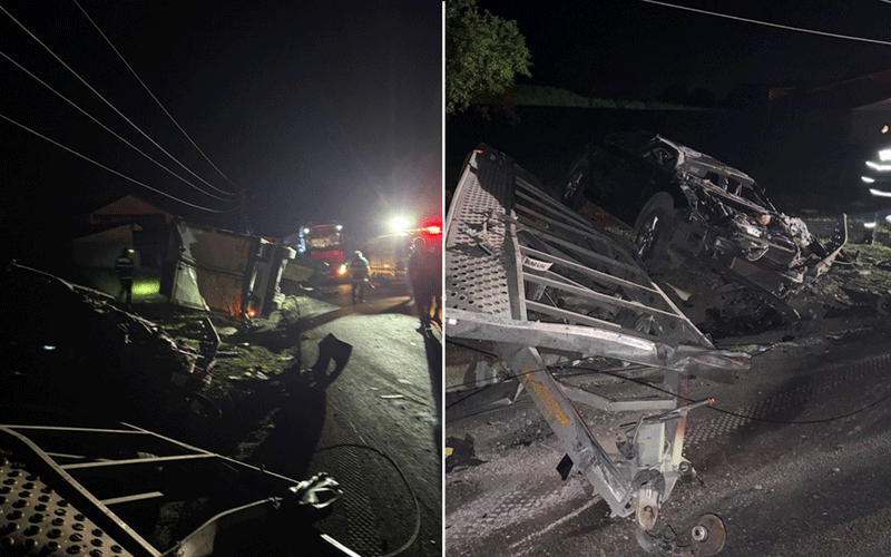 Nighttime crash scene with an overturned trailer, damaged vehicle, and responders illuminated by emergency lights on a dark road.