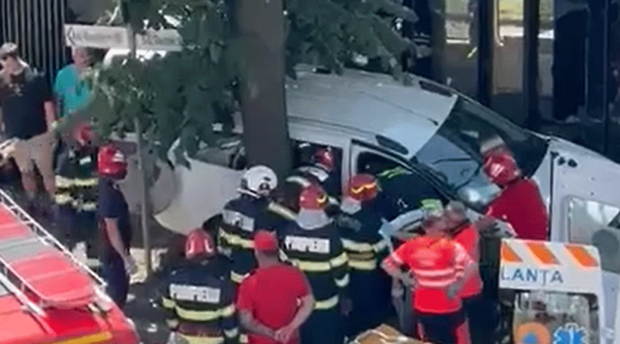 Emergency responders kneel around a white vehicle crashed into a tree as firefighters assist at the scene, with bystanders nearby.