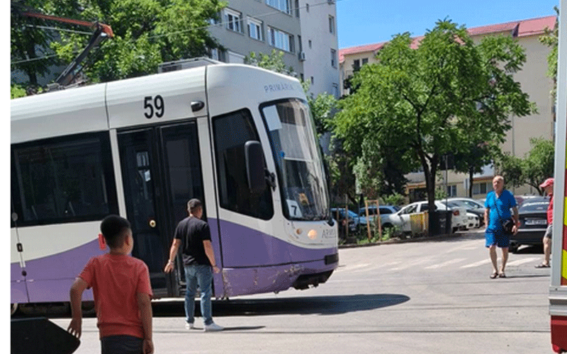 White and purple tram tilting on a sunny city street, with pedestrians nearby and trees and apartment buildings in the background.