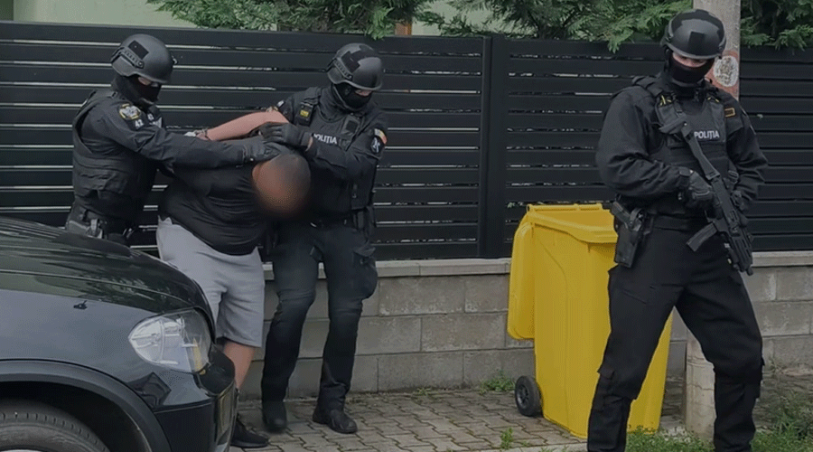 Three heavily armed police officers restrain a man against a low wall beside a black car and a yellow trash bin nearby.