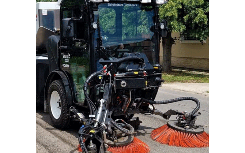 Front view of a street sweeper with orange bristle brushes cleaning a street on a sunny day.