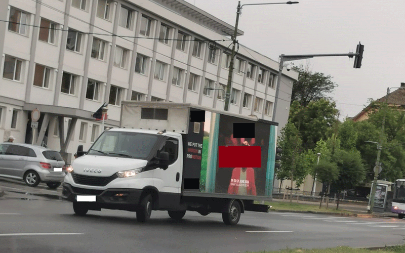 banner-publicitar - Sursa de Vest White delivery truck with a large advertising trailer driving through a city street in front of a multi-story building.