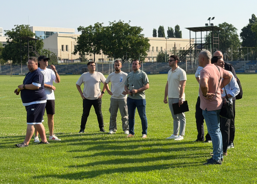 Group of men standing in a circle on a grassy sports field, chatting during a practice or meeting, with bleachers and buildings in the background.