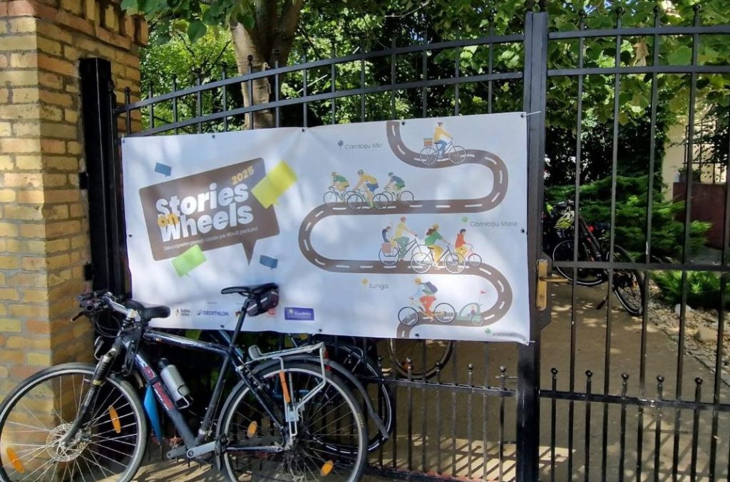 Banner reading 'Stories on Wheels' with a winding bike path illustration on a black iron gate; several bicycles parked in front on a sunny day.