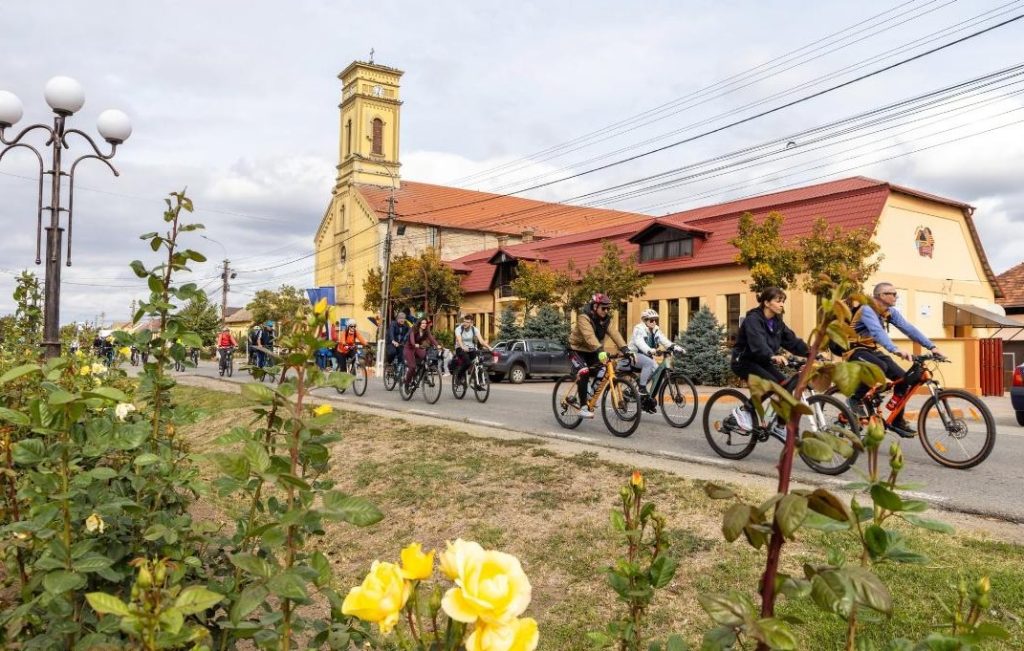 Group of cyclists ride along a town street past a yellow church with red roof, seen through a foreground bed of yellow roses and greenery.