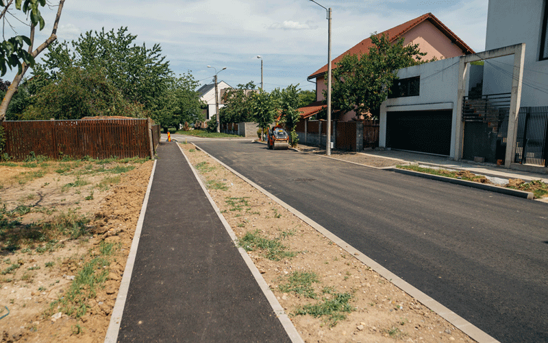 blascovici - Sursa de Vest Suburban street with a paved road on the right and a parallel pedestrian/bike path on the left, under a clear sky.