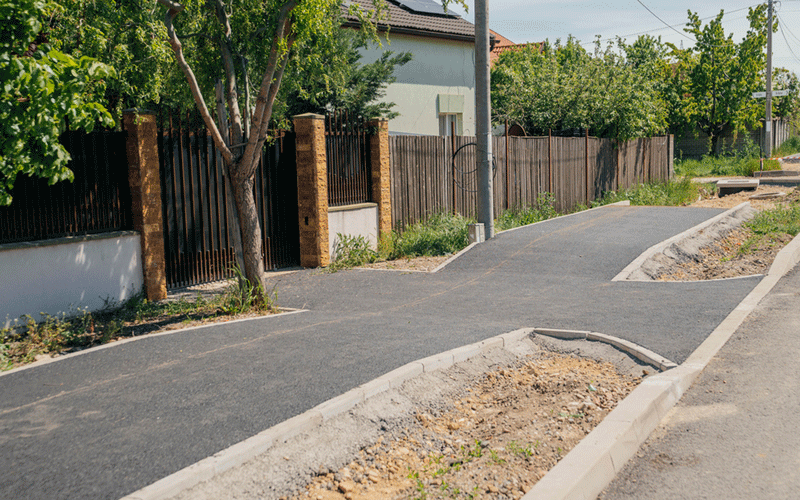 blascovici2 - Sursa de Vest New paved sidewalk along a residential street with a dirt median, wooden fences, and trees; ongoing construction and bare soil patches nearby.