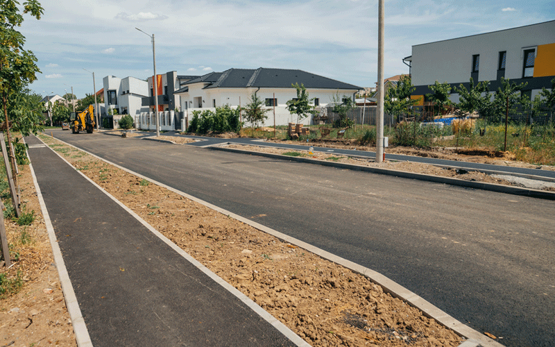 Street under construction in a new residential neighborhood; an excavator on the left and modern houses ahead.