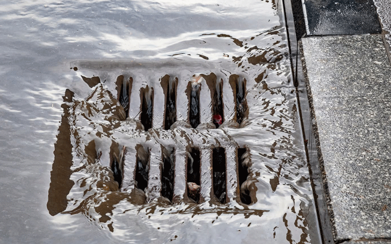 Storm drain with muddy water spilling over a metal grate onto a sidewalk edge, urban setting.